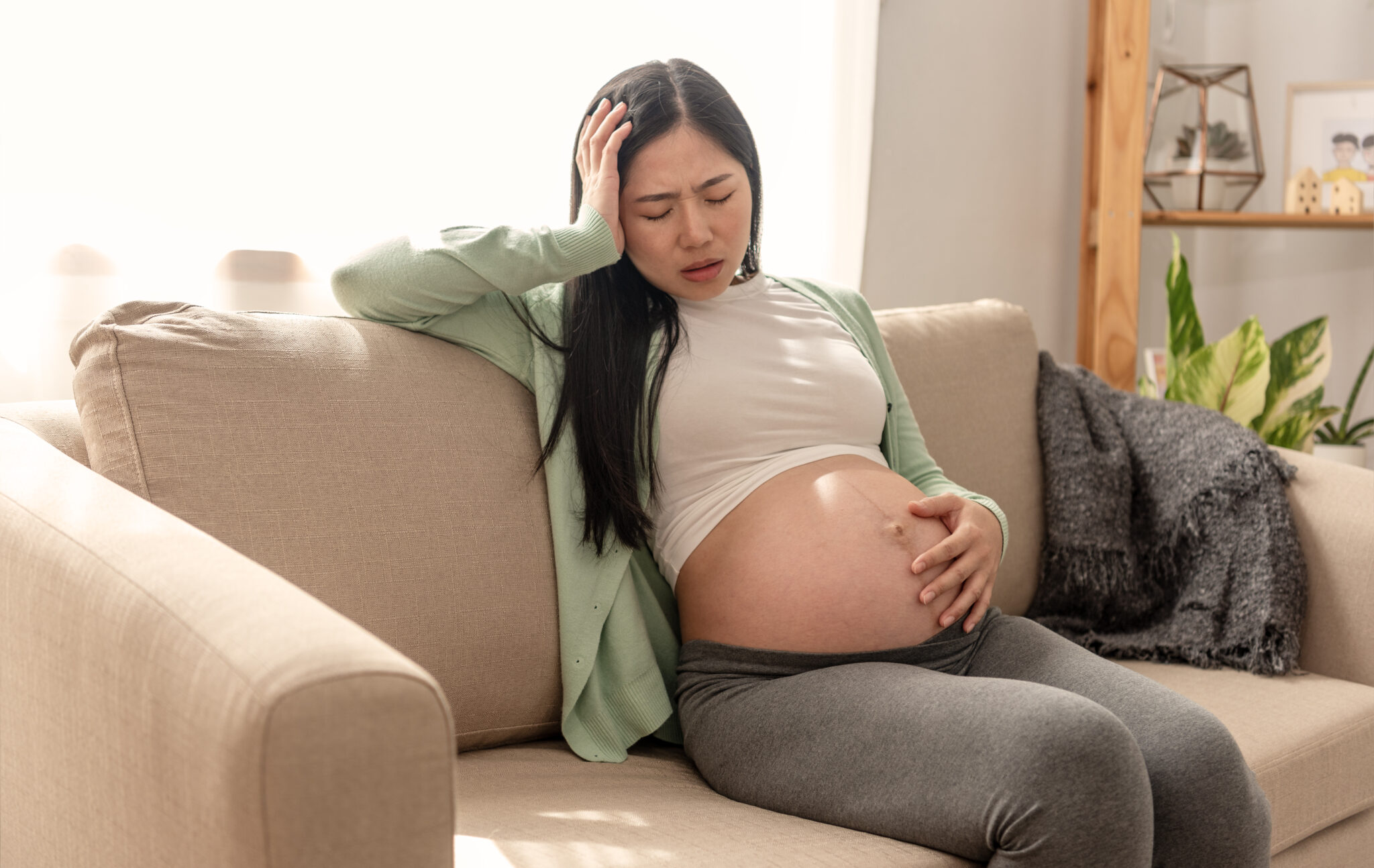 Asian young pregnant woman suffering belly ache sitting on a couch in the living room at home.