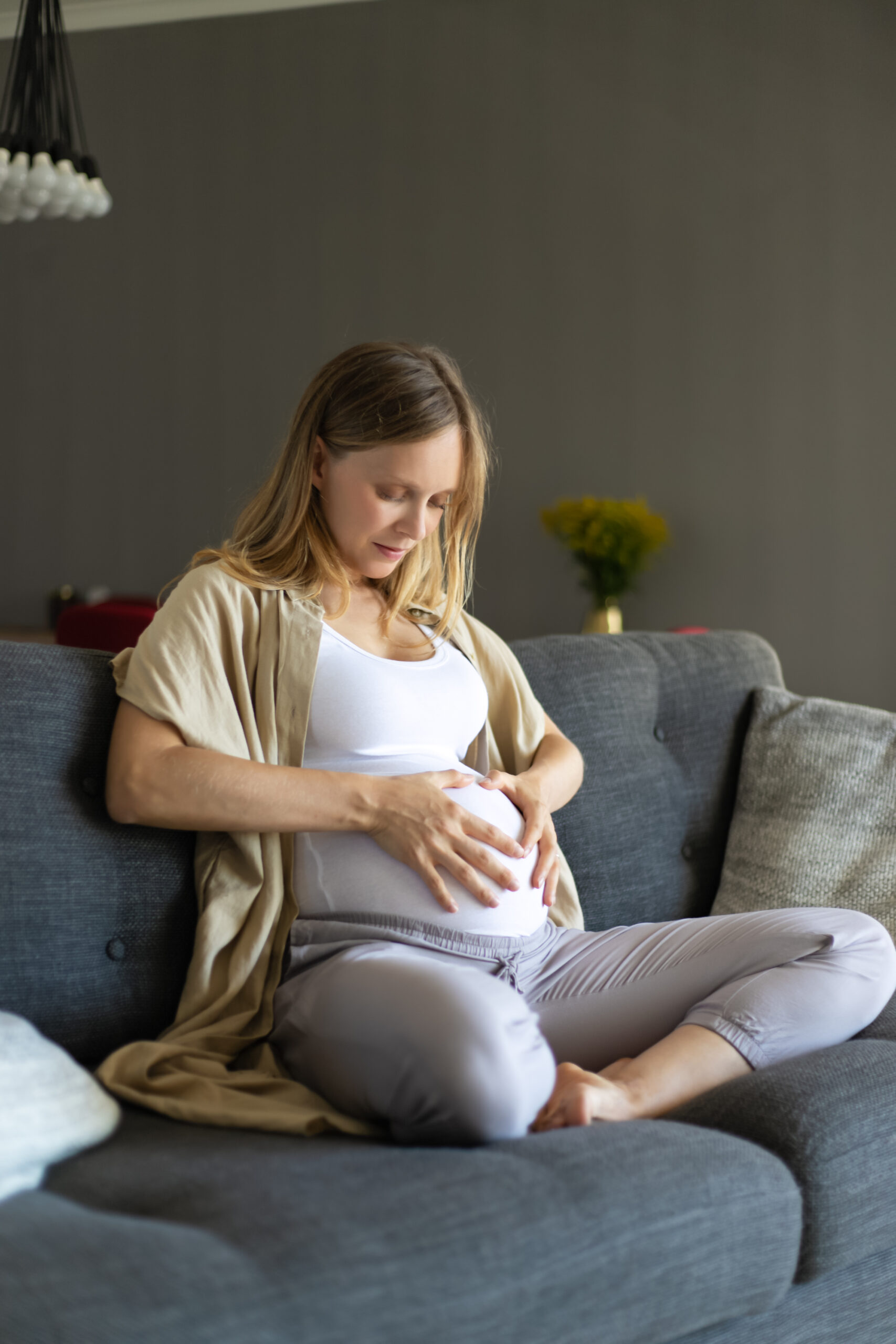 Serene focused expectant mother sitting on couch, touching belly, feeling moving inside stomach. Pregnant young woman spending leisure time at home. Enjoying pregnancy concept