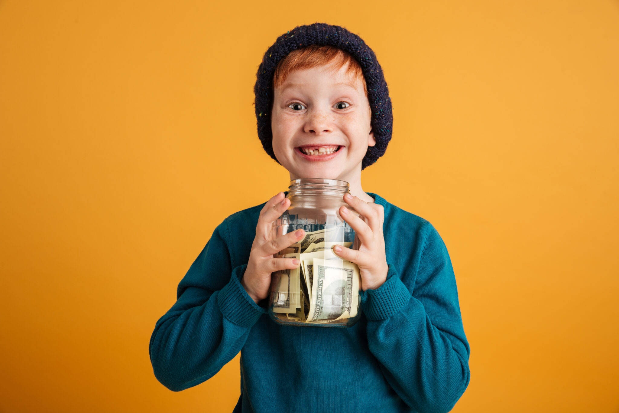 Photo of funny little redhead boy with freckles standing isolated over yellow background wearing warm hat. Looking camera holding money.