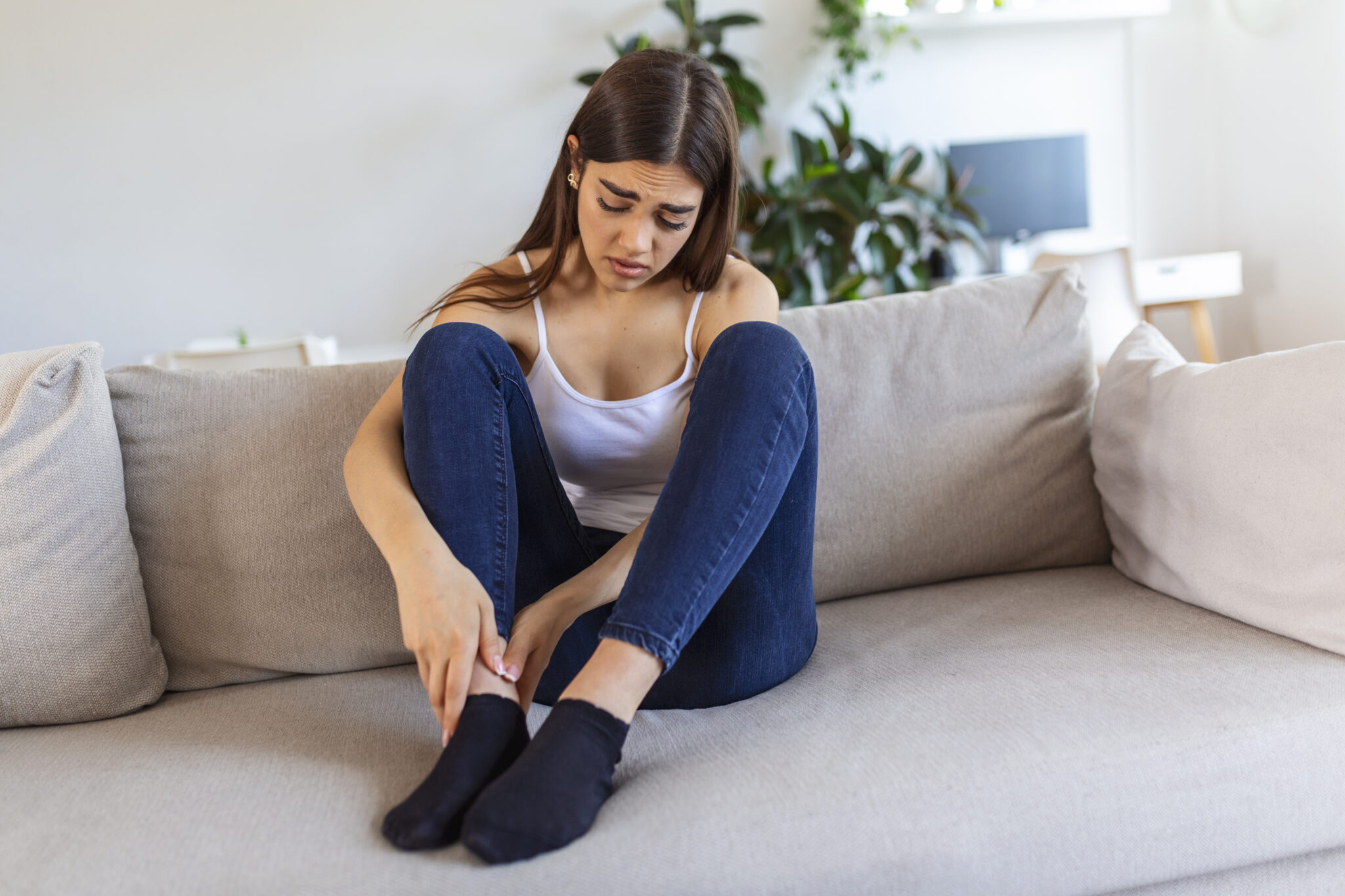 Hands massaging swollen foot while sitting on sofa during the day at home. Photo of Young Caucasian woman suffering from pain in leg. Woman massaging her legs after all day at work in office