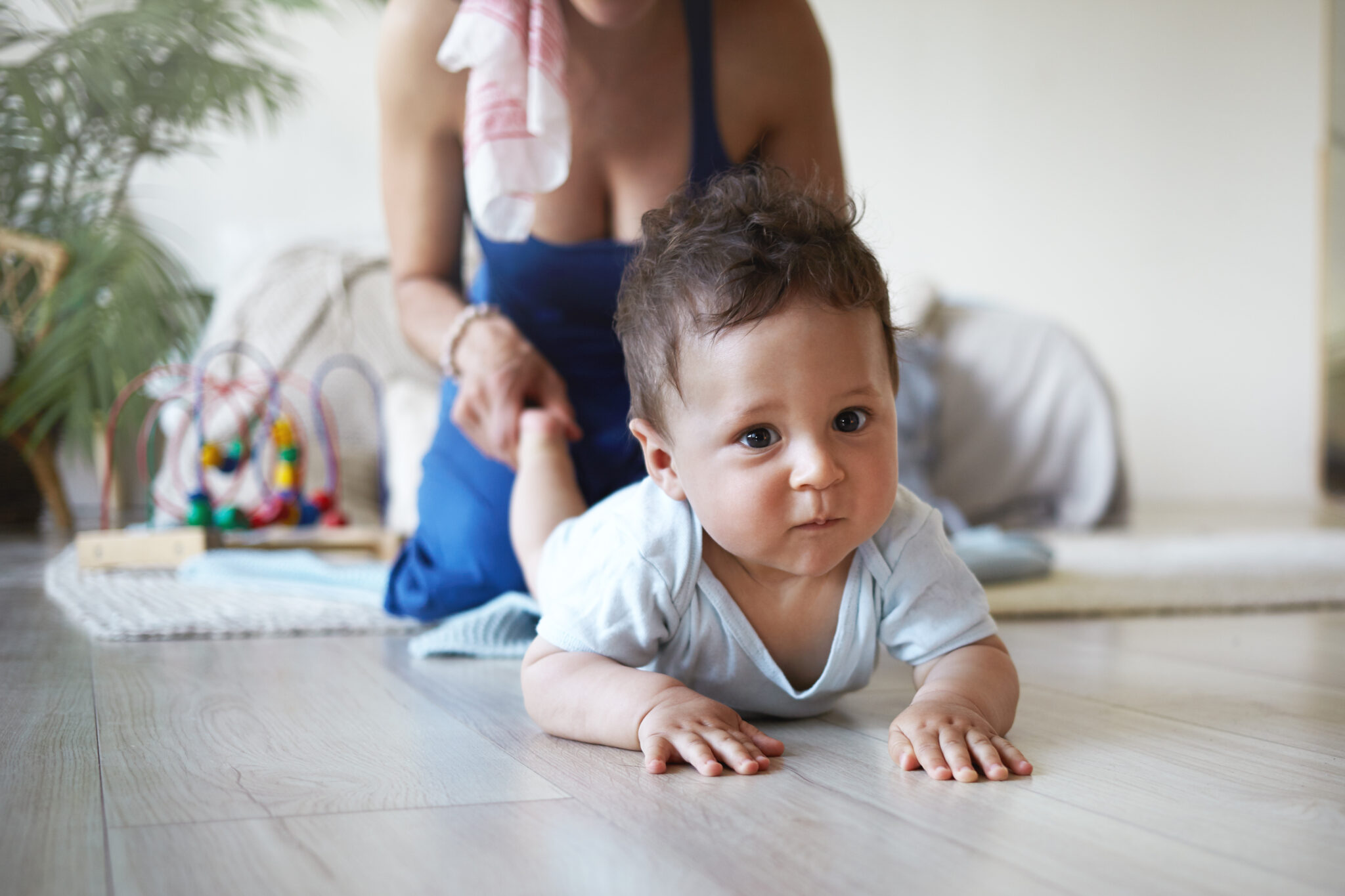 Cropped shot of positive young mixed race female with charming smile holding toes of her baby son while playing with him on floor at home. Parenting, childhood, maternity and family bonding concept