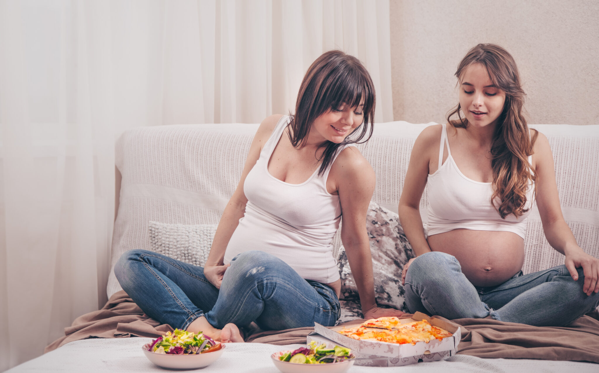 Maternity concept, two pregnant women eating pizza and salad at home on the sofa, friendship and healthy eating