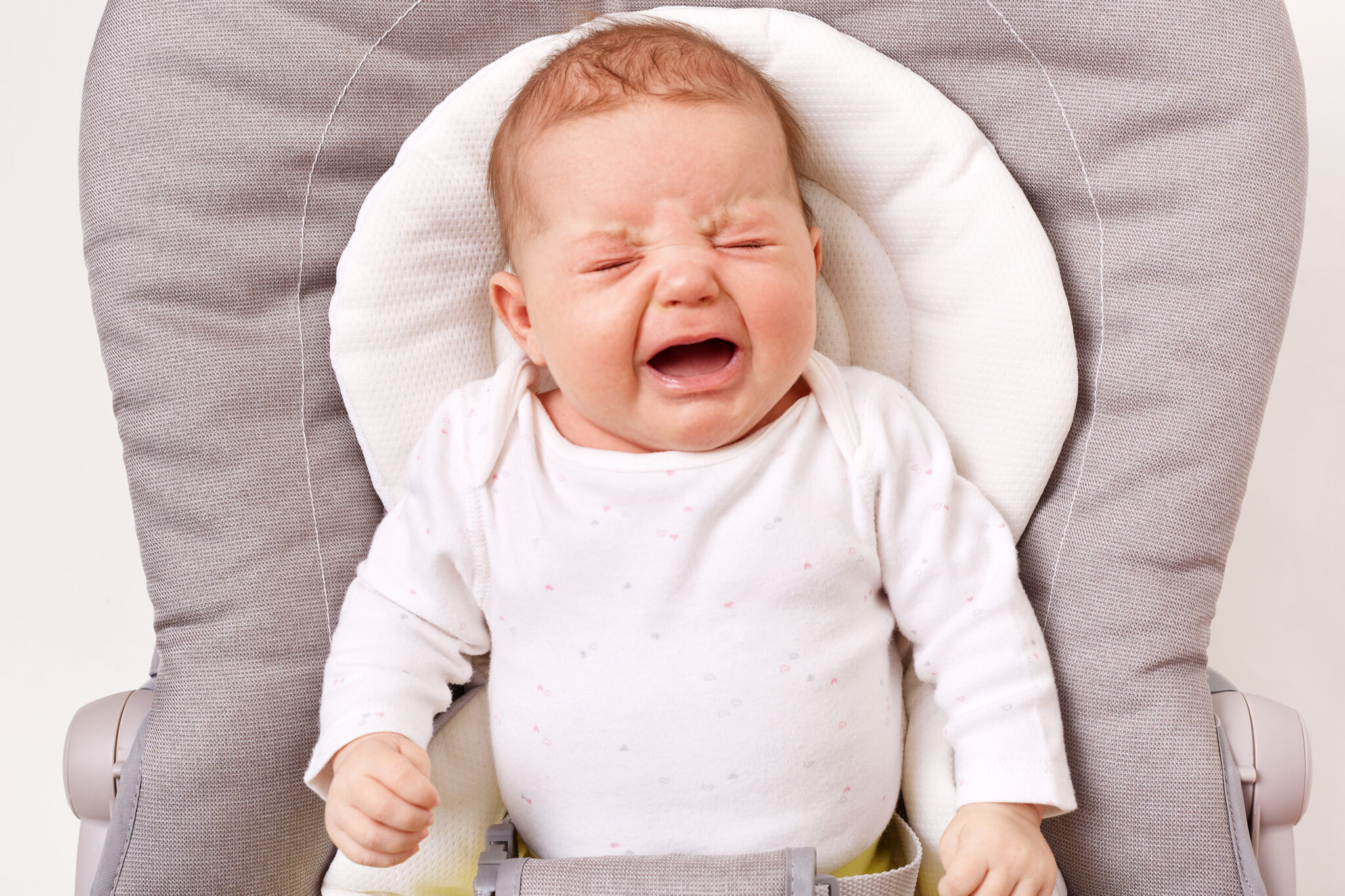 Unhappy little infant girl or boy crying in bouncer chair, being hungry, wants to sleep, needs mother's attention, wearing white shirt, keeps eyes closed, hands in tiny fists.