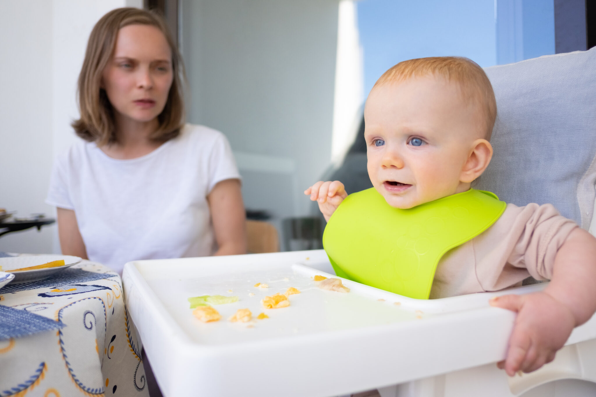 Young mom watching cheerful cute baby sitting in high chair with messy in tray. First solid food or child care at home concept