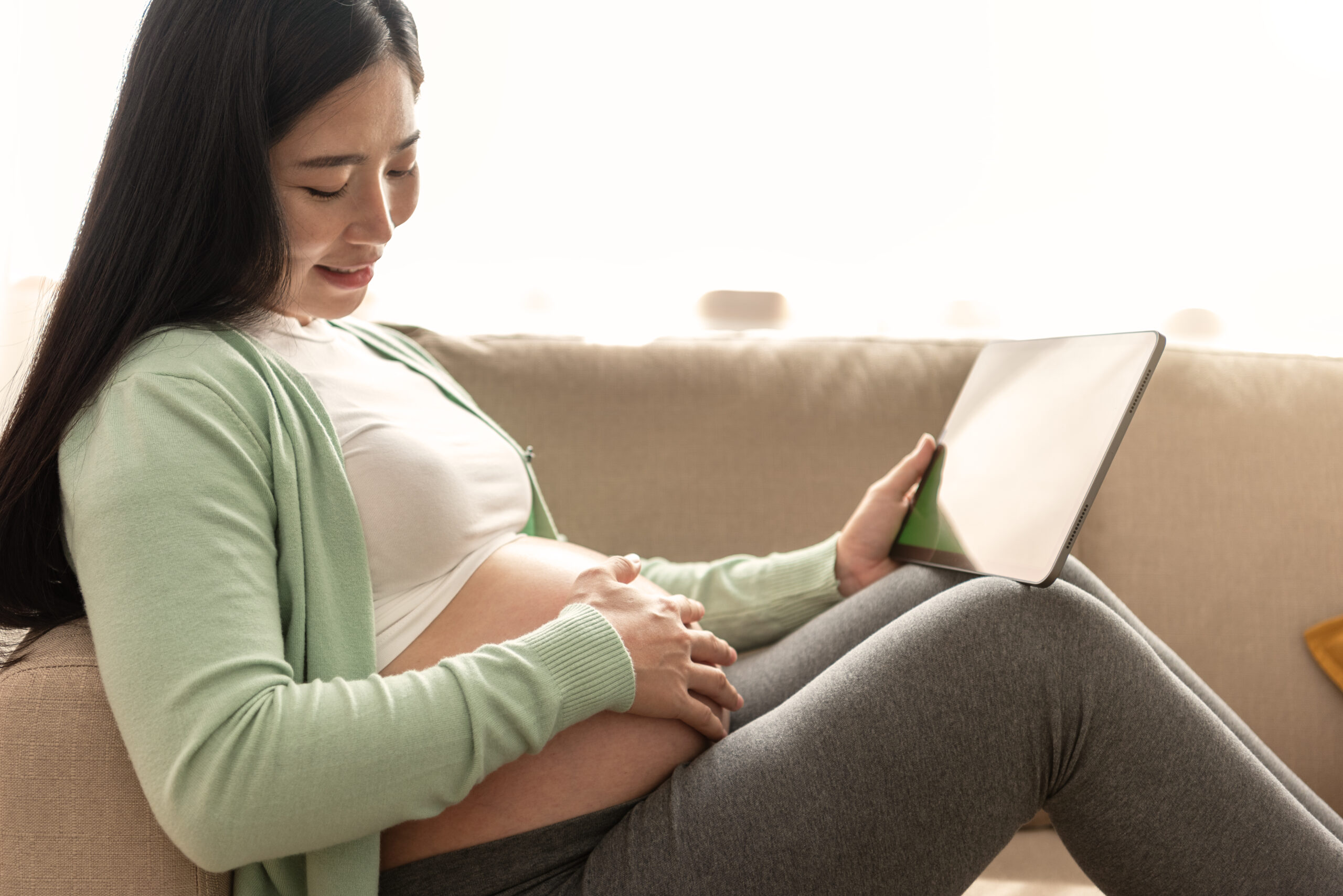 Young asian pregnant woman hands fondle on belly and holding green chroma key screen on tablet sitting on sofa at home. doctor online.