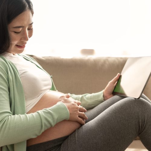 Young asian pregnant woman hands fondle on belly and holding green chroma key screen on tablet sitting on sofa at home. doctor online.